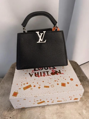 Iouis Vuitton handväska  - Detta är en handväska i svart, strukturerat läder med en stilren och lyxig estetik. Väskan har en trapetsformad silhuett med en framträdande flikstängning. Den mest iögonfallande detaljen är en stor, silverfärgad metallogo i form av två sammanflätade bokstäver som fungerar som ett dekorativt lås på framsidan.