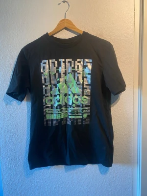 Svart Adidas t-shirt med tryck - Säljer en svart t-shirt från Adidas med ett stort grafiskt tryck i silver och grönt på bröstet. Klassisk rund hals och korta ärmar. Tillverkad i mjuk bomull som är skön att bära. Perfekt för dig som gillar sportig streetstyle.