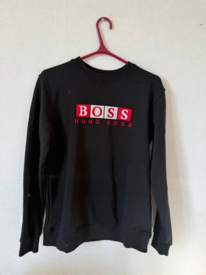 Svart sweatshirt från Hugo Boss - Svart sweatshirt från Hugo Boss med stor broderad logga i rött och vitt på bröstet. Klassisk rund halsringning och långa ärmar. Perfekt för en clean och stilren look.