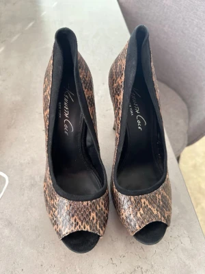 Leopardmönstrade pumps Kenneth Cole - Coola pumps från Kenneth Cole med leopardmönster i brunt och svart. Skorna har peep toe och klädd klack, samt svarta kanter runt öppningen. Materialet har en struktur som liknar ormskinn för en extra edgy känsla.