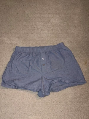 Blå boxerkalsonger/ pyjamas short med knappgylf - Säljer ett par klassiska blå boxerkalsonger/ pyjamasshorts i tunn bomull med två knappar i gylfen. De har en avslappnad passform och resår i midjan för extra komfort. Perfekta för dig som gillar enkel och stilren design.