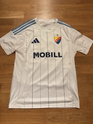 Djurgårdens bortamatch tröja från säsongen 2024 - Säljer en vit Djurgården matchtröja från Adidas med tunna blå ränder, klubbmärke på bröstet och sponsortryck. Tröjan har ljusblå ränder på axlarna, namn och nummer 21 Lucas Bergvall på ryggen. Tillverkad i lätt och ventilerande material, perfekt för fotboll. Köpt på Djurgårds butiken. Den har även en signering av lucas bergvall som har börjat försvinna lite