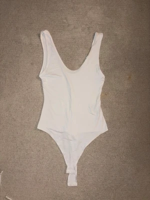 Vit basic body med djup ringning - En enkel och stilren vit body med breda axelband och djup rundad ringning fram. Bodyn har klassisk skärning och knäppning i grenen. Perfekt att matcha med jeans eller kjol för en clean look.
