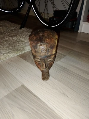 Träskulptur med ansikte - Unik träskulptur i brunt trä som föreställer ett stiliserat ansikte med inhuggna detaljer och geometriskt mönster på pannan. Skulpturen har en avlång form och är handgjord, perfekt som dekoration eller samlarobjekt.