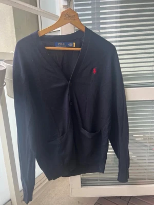 Mörkblå cardigan från Polo Ralph Lauren - Stilren mörkblå cardigan med knappar från Polo Ralph Lauren. Tröjan har två fickor framtill, V-ringning och den klassiska röda logotypen broderad på bröstet. Tillverkad i mjukt material som passar perfekt till lager-på-lager.