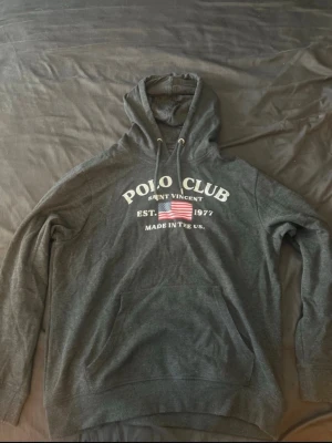 Mörkgrå hoodie Polo Club - Mörkgrå hoodie från Polo Club med tryck framtill som säger 'Saint Vincent, Est. 1977, Made in the US.' och en amerikansk flagga. Klassisk känguruficka och justerbar huva med snören. Perfekt för en avslappnad och sportig stil.