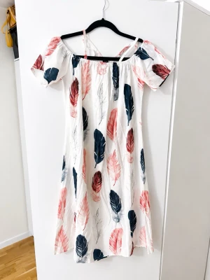 Vit offshoulder klänning med fjädermönster - Luftig vit klänning med offshoulder och smala axelband. Klänningen har ett färgglatt fjädermönster i rosa, svart och rött. Den är knälång och har korta ärmar. Perfekt för varma dagar och ger en avslappnad, bohemisk känsla.