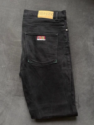 Svarta jeans från Kenzo - Svarta jeans från Kenzo med klassisk femficksdesign och Kenzo-lapp bak i midjan. Jeansen har en färgglad patch på bakfickan och gröna detaljer vid fickorna. Tillverkade i denim med rak passform. Observera hål mellan benen enl bilder och slitet vid ena fickan. 
