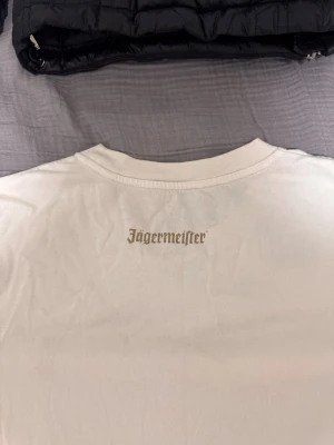 Vit Jägermeister t-shirt med tryck - Vit t-shirt med Jägermeister-tryck i nacken och ett litet J på bröstet. Klassisk rund halsringning och korta ärmar. Perfekt för dig som gillar stilrena plagg med en twist.