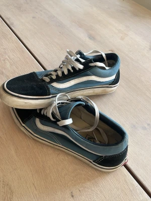 Blå och svarta Vans Old Skool sneakers - Klassiska Vans Old Skool sneakers i blått och svart med vit sula och vit sidosnirk. Skorna har låga skaft, vita skosnören och är tillverkade i en mix av mocka och canvas. Perfekta för en avslappnad streetstil.