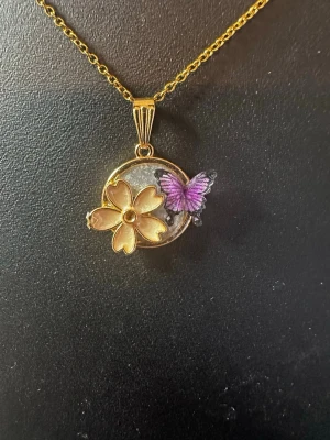 Guldigt halsband med blomma och fjäril - Unikt halsband med en rund guldpläterad berlock som pryds av en guldfärgad blomma och en lila fjäril. Kedjan är tunn och elegant, perfekt för att ge en söt och färgglad touch till din stil. Materialet ser ut att vara metall med emaljdetaljer.