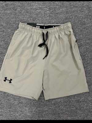 Beige shorts med snörning Under Armour - Snygga beige shorts från Under Armour med svart snörning i midjan och logga på vänster ben. Perfekta för träning eller chill dagar. Lätta och bekväma med elastisk midja och sidofickor.