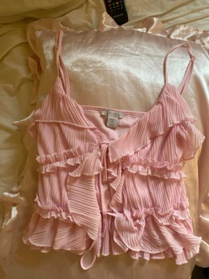 Rosa volangtop med tunna axelband - Supersöt rosa topp med tunna axelband och volangdetaljer. Toppen har plisserat tyg och flera lager volanger framtill samt knytning i mitten. Perfekt för dig som gillar en romantisk och lekfull stil. Materialet är lätt och luftigt.