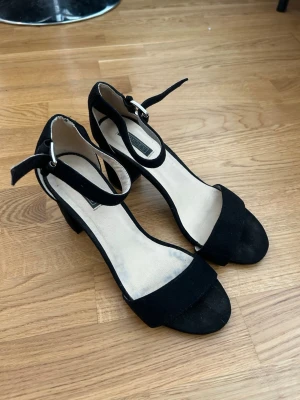 Svarta pumps med ankelrem - Stilrena svarta pumps med öppen tå och bred klack. Skorna har en snygg ankelrem med spänne och är klädda i mockaliknande material. Perfekta för att lyfta din outfit med en elegant touch.