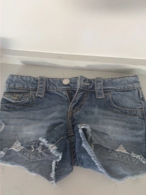 Blå jeansshorts med slitningar - Säljer ett par blå jeansshorts med råa kanter och slitna detaljer framtill. Shortsen har fem fickor, bälteshällor och knappstängning. Perfekta för varma dagar och ger en avslappnad look. Storlek 146. 