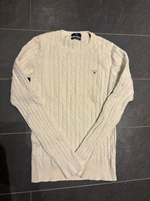 Vit stickad tröja från Gant - Kabelstickad vit tröja från Gant med rund halsringning och diskret logga broderad på bröstet. Tröjan har långa ärmar och ribbade muddar vid ärmslut och nederkant. Perfekt för dig som gillar klassisk och stilren design.