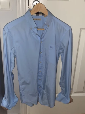 Ljusblå skjorta från Burberry  - Säljer en klassisk ljusblå skjorta från Burberry Brit. Skjortan har button-down krage, långa ärmar och diskret logga broderad på bröstet. Tillverkad i mjuk bomull och stängs med knappar framtill. Perfekt för en clean och stilren look. Kom med bud