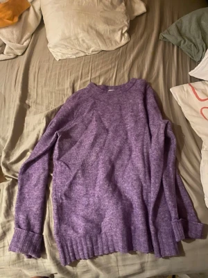 Lila stickad tröja med rund hals - Mysig lila stickad tröja med rund hals och långa ärmar. Tröjan har ribbstickade muddar vid ärmslut och nederkant, vilket ger en avslappnad och trendig look. Perfekt för kalla dagar och enkel att matcha med jeans eller kjol.