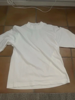 Uniqlo T-shirt - Säljer en stilren vit t-shirt från Uniqlo med högre krage (crew T-shirt). T-shirten har en baggy passform med korta ärmar. Perfekt basplagg för alla tillfällen och enkel att matcha med olika outfits. Det är AlRism tyg, vilket är bra på sommaren då den bland annat fångar upp svett. Inga skavanker.
