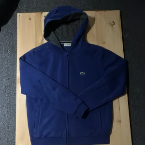 Marinblå hoodie från Lacoste - Säljer en marinblå hoodie från Lacoste med dragkedja och klassisk krokodillogga på bröstet. Tröjan har huva med grått foder, långa ärmar och två fickor framtill. Perfekt för dig som gillar stilren och sportig look. Säljer den för den va alldeles för liten när jag köpte den så har bara luggit i garderoben så köp gärna vill bli av med den kom gärna med bud.👍