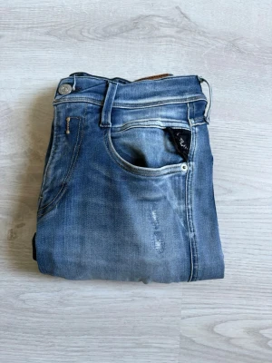 Blå slitna jeans med tvättade detaljer - Säljer ett par blå jeans med slitningar och tvättade detaljer. Jeansen har klassisk femficksmodell, smal passform och snygga slitningar på benen för en trendig look. Perfekta för dig som gillar en avslappnad och cool stil.