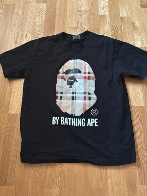 Svart t-shirt med rutigt tryck - Säljer en svart t-shirt med det klassiska apansiktet i stort rutigt mönster på bröstet. T-shirten har rund hals och korta ärmar. Perfekt för dig som gillar streetwear och vill sticka ut.