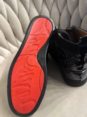 Svarta sneakers med nitar och röd sula - Unika svarta sneakers med glansiga detaljer och svarta nitar på sidorna. Skorna har en ikonisk röd sula och snörning framtill. Materialmix av lack och textil ger en cool kontrast och exklusiv känsla. Perfekta för dig som vill sticka ut med din stil.