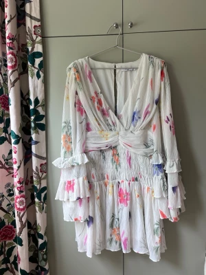 Blommig kortklänning från NA-KD - Supersöt kortklänning från NA-KD i vitt med blommigt mönster i flera färger. Klänningen har v-ringning, volangdetaljer på ärmarna och midjan samt långa ärmar. Perfekt för sommarens alla tillfällen!