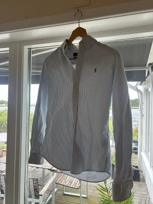 Randig skjorta från Polo Ralph Lauren - Klassisk skjorta från Polo Ralph Lauren med smala blå och vita ränder. Skjortan har knappslå, button-down krage och den ikoniska broderade loggan på bröstet. Tillverkad i bomull och har långa ärmar med manschetter.
