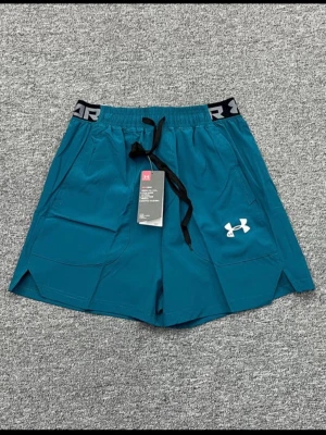 Under Armour träningsset blågrön - Snyggt träningsset från Under Armour med blågröna shorts och matchande t-shirt. Shortsen har elastisk midja med snörning och logga, samt fickor. T-shirten har diskret mönster och logga på bröstet. Perfekt för gym eller löpning.