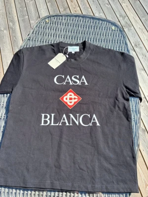 Svart t-shirt från Casablanca - Snygg svart t-shirt från Casablanca med stort vitt tryck 'CASA BLANCA' och en röd logga på bröstet. Klassisk rund hals och korta ärmar. Perfekt för dig som gillar stilrena plagg med statement-tryck.