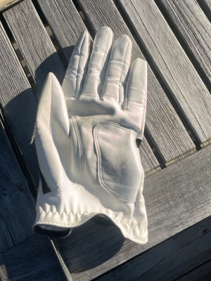 Vit golfhandske från FootJoy - Snygg vit golfhandske från FootJoy med svarta detaljer och justerbar kardborreknäppning. Handsken har perforeringar för extra ventilation och är tillverkad i mjukt skinn för bra grepp och komfort på golfbanan.