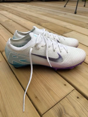 Vita Nike Mercurial fotbollsskor - Säljer ett par vita Nike Mercurial fotbollsskor med lila och blå detaljer. Skorna har en låg profil, snörning och en syntetisk ovandel. Sulan är i metallic blå och lila med fasta dobbar för bra grepp på planen. Perfekta för dig som vill sticka ut på fotbollsplanen!               Säljer för att det är för stora och är använd 2 träningar 