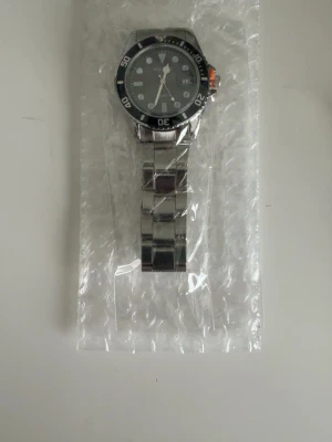 Klassisk silverfärgad armbandsklocka - Stilren armbandsklocka med svart urtavla och silverfärgad länk i metall. Klockan har tydliga siffror och markeringar på boetten samt en orange detalj på sidan. Perfekt accessoar för en modern look.