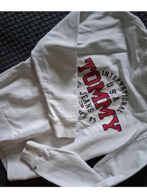 Vit sweatshirt från Tommy Jeans - Vit sweatshirt från Tommy Jeans med stort rött och svart tryck på bröstet där det står 'TOMMY INTERNATIONAL U.S.A JEANS'. Tröjan har rund halsringning, ribbade muddar och är i regular fit. Perfekt för en clean och sportig look.