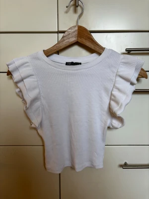 Vit ribbad topp i bomull från Zara med volangärmar, storlek S - Säljer en vit ribbad bomullstopp med volangärmar. Toppen har rund halsringning och är något croppad i modellen. Storlek S - passar även XS