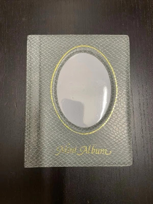 Grått mini fotoalbum - Säljer ett mini fotoalbum i grått med ormskinnsmönster och gulddetaljer. Omslaget har ett ovalt fönster för egen bild och texten 'Mini Album' i guld. Insidan har 12 vita dubbelsidiga plastfickor för foton. Perfekt för att samla minnen eller ge bort som present.