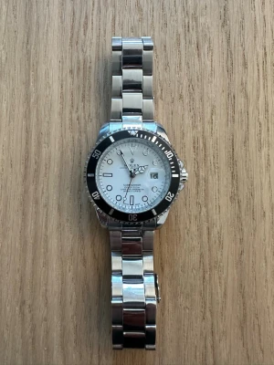 Submariner silverklocka - Stilren Submariner med vit urtavla, svart vridbar ring och datumfönster. Klockan har en klassisk länk i silverfärgat stål och tydliga indexmarkeringar. Perfekt för dig som gillar tidlös design och exklusiv känsla. Helt ny. Aldrig använd.