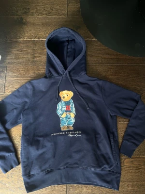 Polo bear hoodie  - Säljer en marinblå hoodie från Ralph Lauren med den ikoniska Polo Bear-trycket på bröstet. Hoodien har huva med dragsko och ribbade muddar. Perfekt för dig som gillar streetwear och vill ha en klassisk look.