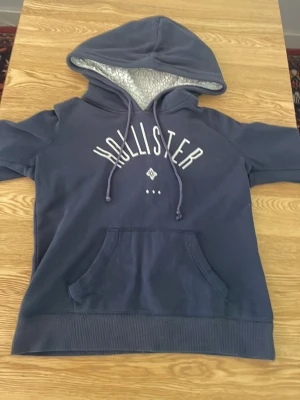 Blå Hollister hoodie med huva - Mysig blå hoodie från Hollister med stor huva och vitt foder. Hoodien har korta ärmar, magficka och dragsko i huvan. Hollister-logga i vitt framtill. Perfekt för chill dagar och streetwear-stil.