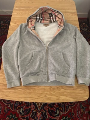 Grå hoodie med Burberry-mönstrad huva - Säljer en grå zip-hoodie från Burberry med den klassiska rutiga mönstringen i huvan. Hoodien har två fickor framtill, ribbade muddar och dragkedja. Perfekt för dig som vill ha en stilren och lyxig vardagslook.