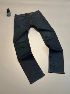Mörkblå jeans från Replay - Säljer ett par mörkblå jeans från Replay med klassisk femficksmodell och raka ben. Jeansen har kontrastsömmar och detaljerade bakfickor med dragkedja. Tillverkade i slitstark denim, perfekta för en avslappnad och stilren look.