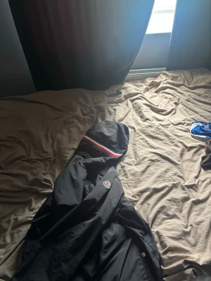Svart Moncler vindjacka med huva - Snygg svart vindjacka från Moncler med huva och klassisk logotyp på bröstet. Jackan har en röd och vit rand på huvan som ger en sportig vibe. Perfekt för blåsiga dagar och enkel att matcha med olika outfits