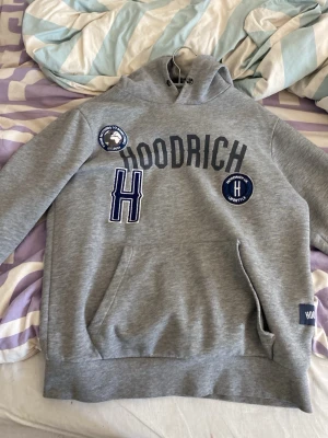Grå hoodie från Hoodrich med tryck - Snygg grå hoodie från Hoodrich med stor logga och tryck på bröstet. Har huva med dragsko och känguruficka framtill. Dekorativa patches och broderad H-detalj i blått och vitt. Perfekt för en streetwear-look.