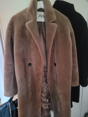 Beige fuskpäls kappa från Zara - Säljer en beige fuskpäls kappa från Zara med bred krage och dubbelknäppning. Kappan har en rak passform och är helfodrad för extra värme. Perfekt för kalla dagar när du vill vara både snygg och varm.