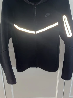 Svart Nike träningsjacka med reflex - Snygg svart träningsjacka från Nike med hel dragkedja och reflekterande detaljer över bröstet, ärmen och längs armen. Jackan har hög krage och är tillverkad i ett stretchigt material som passar perfekt för löpning eller annan träning utomhus.         Hoodie XS Byxor S