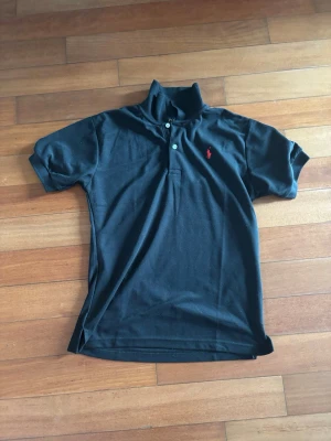 Svart pikétröja med röd logga - Klassisk svart pikétröja med korta ärmar och krage. Tröjan har två knappar framtill och en broderad röd logga på bröstet. Perfekt till jeans eller shorts för en clean och stilren look.