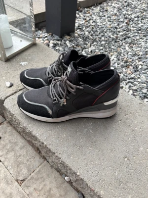 Svarta sneakers från Michael Kors - Snygga svarta sneakers från Michael Kors med grå och vita detaljer samt en diskret röd linje vid hälen. Skorna har snörning, en tjock vit sula och materialmix av textil och mockaimitation. Perfekta för en trendig och sportig look.