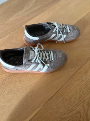 Gråa Adidas sneakers med mocka - Snygga bruna Adidas sneakers i mocka med klassiska vita ränder på sidorna och vita skosnören. Yttersulan är i gummi med en brun ton och har ett mönstrat grepp. Låg modell som passar perfekt till streetwear och avslappnade outfits.