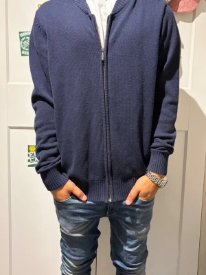 Mörkblå ziptröja i bomull  - Mörkblå ziptröja med ribbade muddar och krage. Tröjan har dragkedja framtill och är tillverkad i mjukt stickat material. Enkel och stilren design som passar till jeans eller chinos.